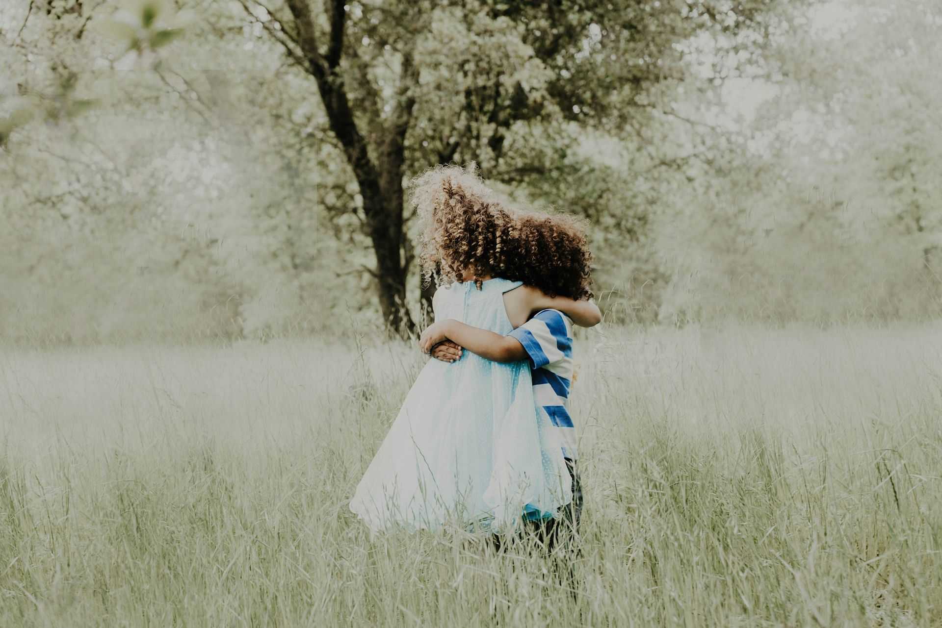 Two children, one in a white dress, hugging in a grassy field with trees in the background.